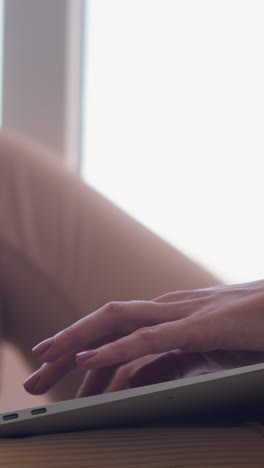 young woman in beige clothes types on keyboard of modern laptop sitting near bright window in light comfortable room at home closeup slow motion