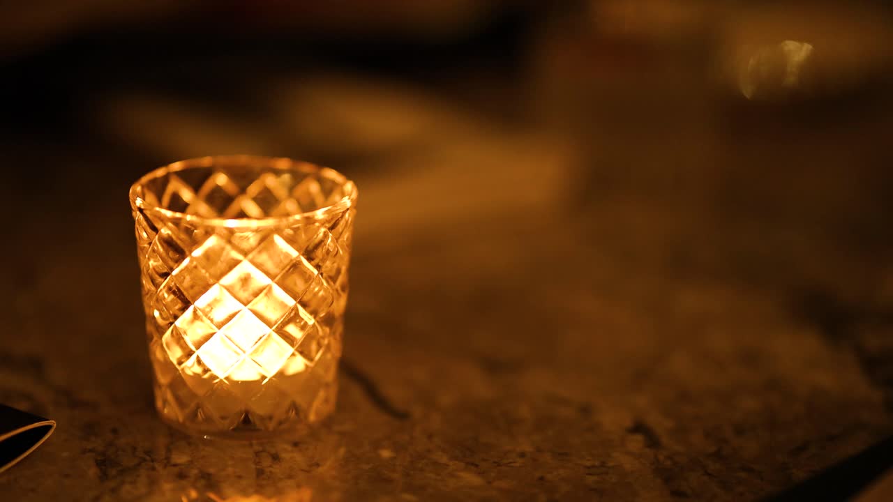 A candle flickers warmly in a patterned glass holder on a dimly lit surface