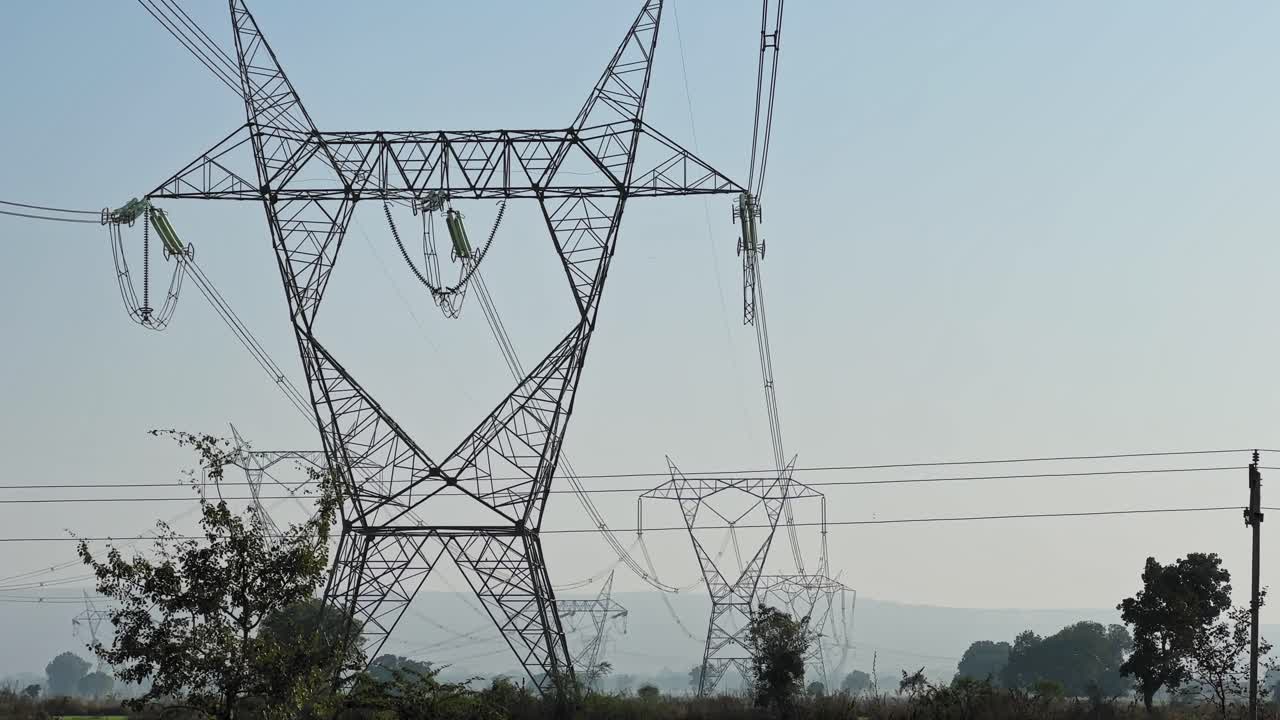 Tilt down shot revealing a series of high voltage transmission towers aligned across green farmland, power lines receding into the distance under soft daylight haze