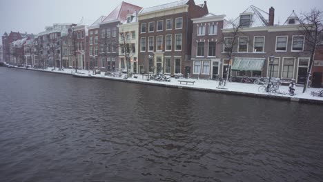 Rhine-river-flowing-through-snowy-Leiden-town-centre-Netherlands,-tilt-up-reveal