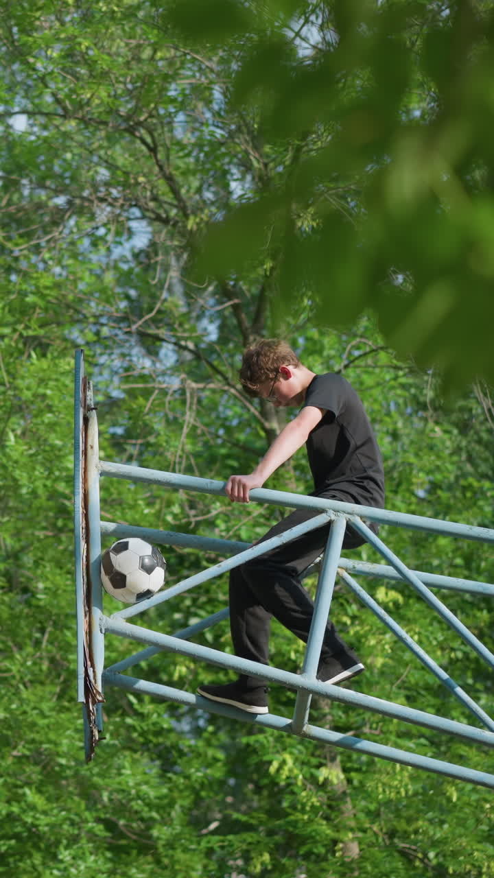 un niño se sienta en un equipo al aire libre de hierro azul con la cabeza inclinada, mientras que una pelota de fútbol está visiblemente atascada en el marco de hierro, con árboles en el fondo