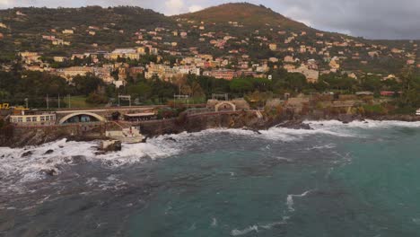 rugged coastline and waves crashing against rocks with genoa city in background, overcast sky, aerial view