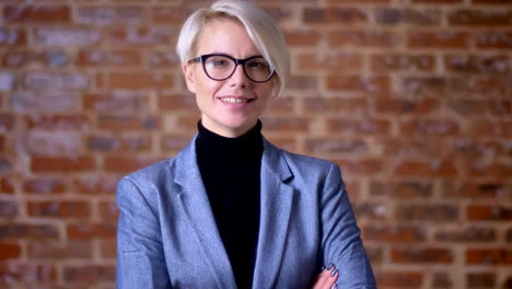 portrait of middle-aged blonde short-haired woman in glasses smiling into camera and nodding on bricken wall background.