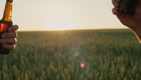 Two-person-clink-bottles-of-beer-against-the-background-of-a-field-of-wheat.-4k-video