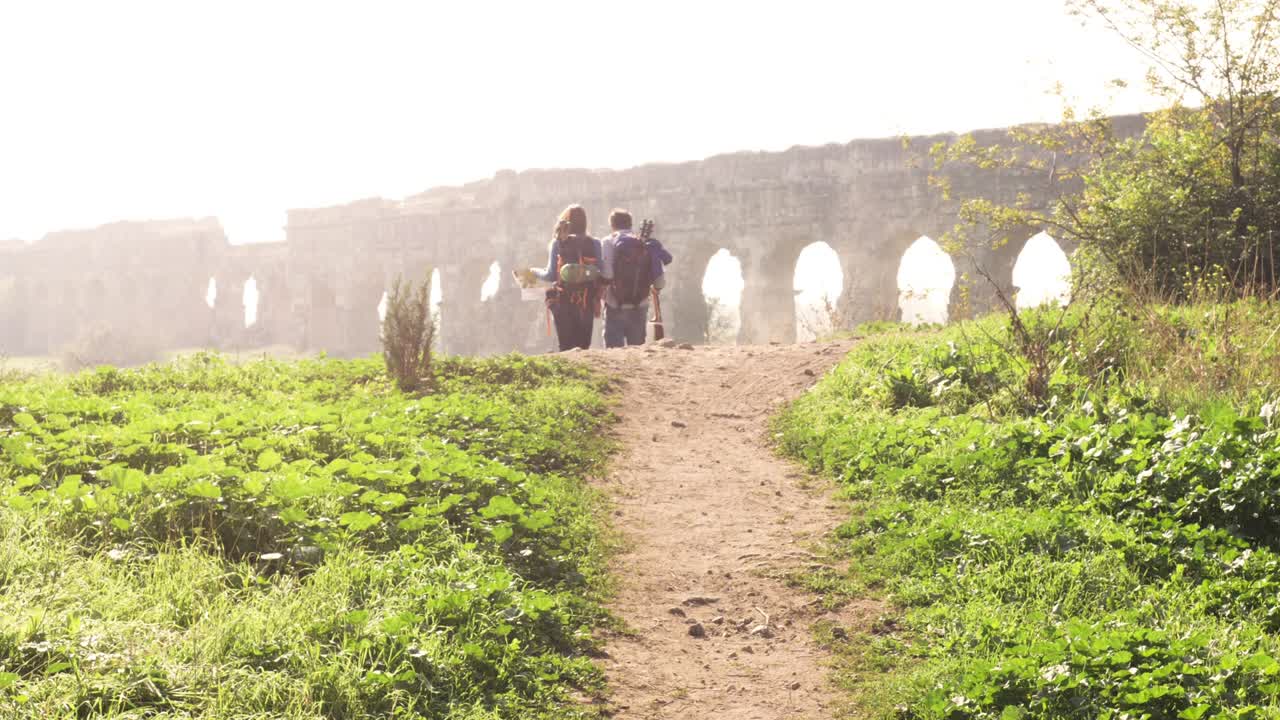 joven pareja encantadora mochileros turistas caminando de la mano hacia el acueducto romano arcos en el parco degli aquedotti ruinas del parque en roma en romántico amanecer brumoso con guitarra y saco de dormir cámara lenta