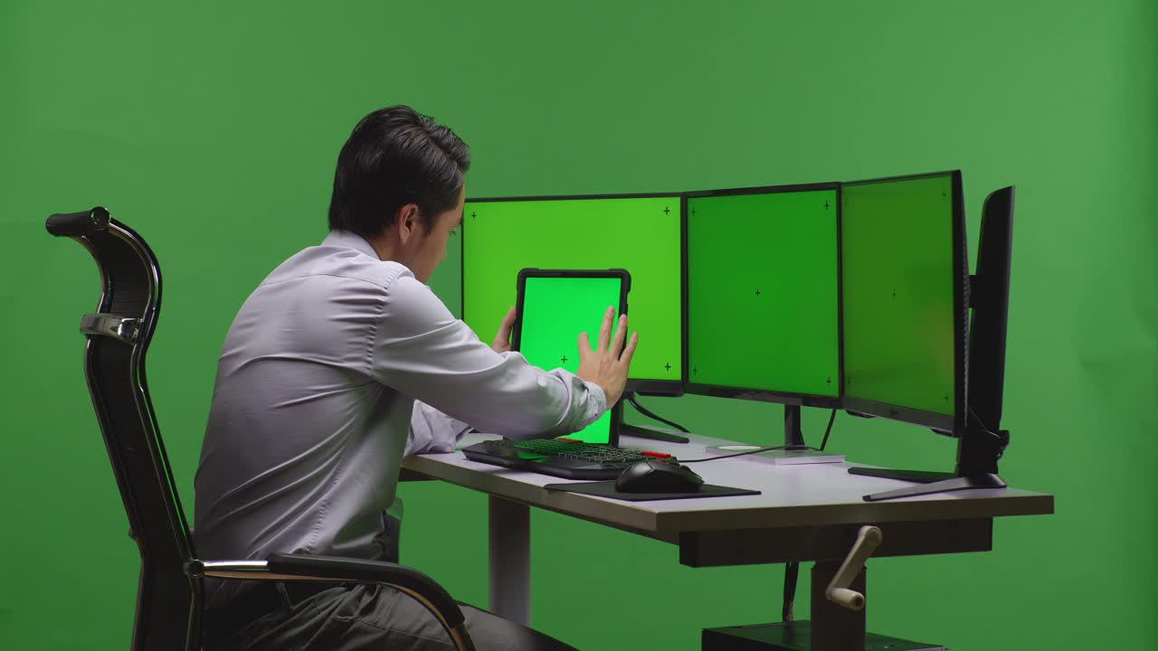 초록색 화면을 가진 여러 모니터 앞의 태블릿에서 일하는 사업가
