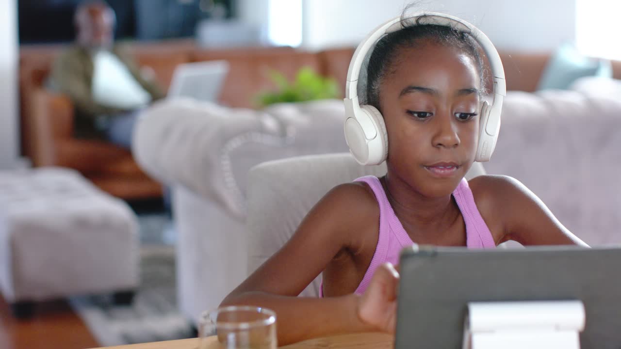 African American girl child wearing tank headset doing online lesson on tablet at living room table