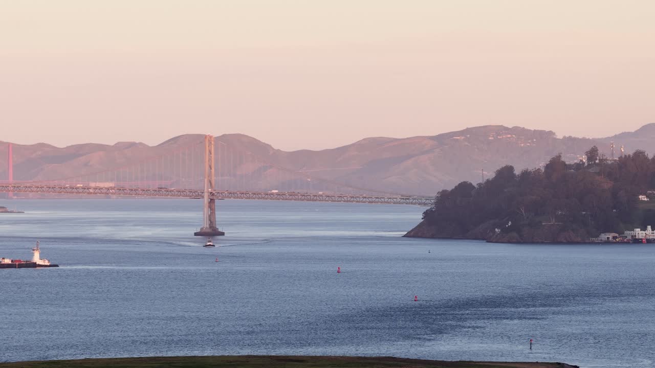 Tomada com teleobjetiva de um rebocador e barcaça navegando pelas águas calmas da Baía de São Francisco ao nascer do sol, com a Ponte da Baía e a Ilha Yerba Buena visíveis através da neblina matinal.