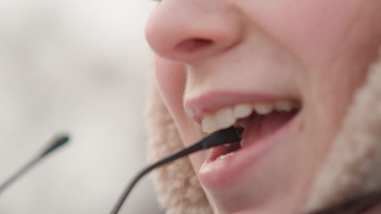 Vista ravvicinata e tagliata del viso inferiore che mostra naso, bocca, mento e denti mentre una donna adulta parla o sorride con le labbra leggermente dischiuse, tenendo un filo sottile vicino alle labbra in una morbida luce invernale esterna.