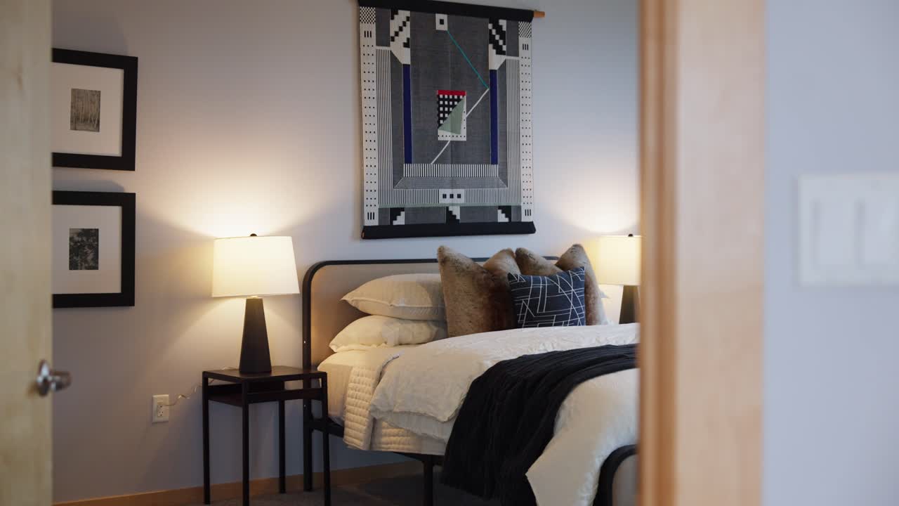 Modern bedroom with warm lighting, a made bed, nightstands, and decorative pillows beneath a patterned wall hanging