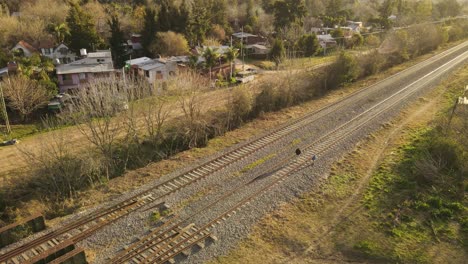 Persona-Adulta-Y-Niño-Caminando-Sobre-Vías-De-Tren-Peligrosas-Durante-La-Puesta-De-Sol,-Vista-Aérea