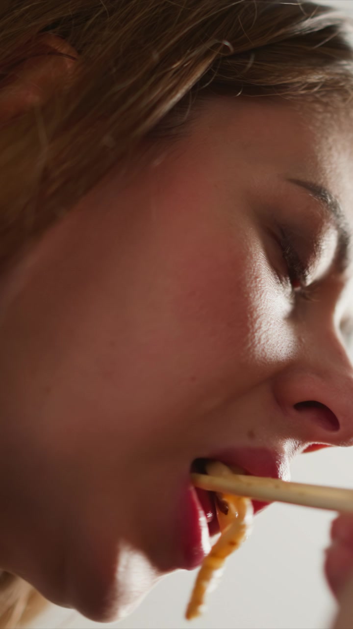 Close up of woman eating noodles focusing on her mouth and facial expressions as woman brings food to her lips. Soft lighting highlights skin texture details against plain background