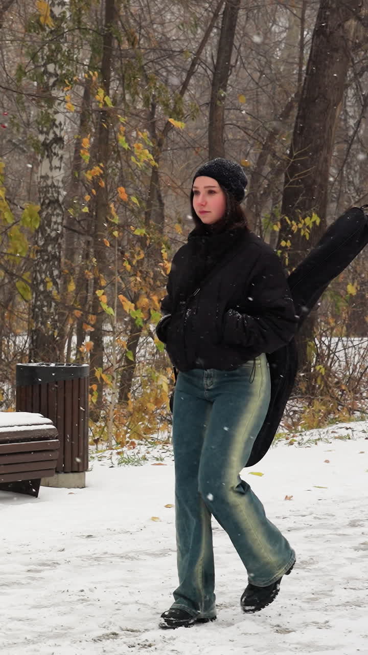 Artista musicale in giacca invernale con le mani in tasca e la chitarra sulla schiena che cammina sotto la nevicata, panchina innevata e alberi secchi sullo sfondo, sereno paesaggio invernale.