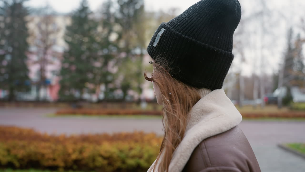 Side view of thoughtful lady in black knit cap and brown shearling jacket walking outdoors on cold overcast day, gentle snowfall adding to serene atmosphere with soft background of park and buildings
