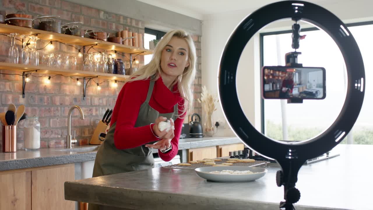 Femme décorant des biscuits avec une poche à douille dans sa cuisine et filmant avec son smartphone pour une démo