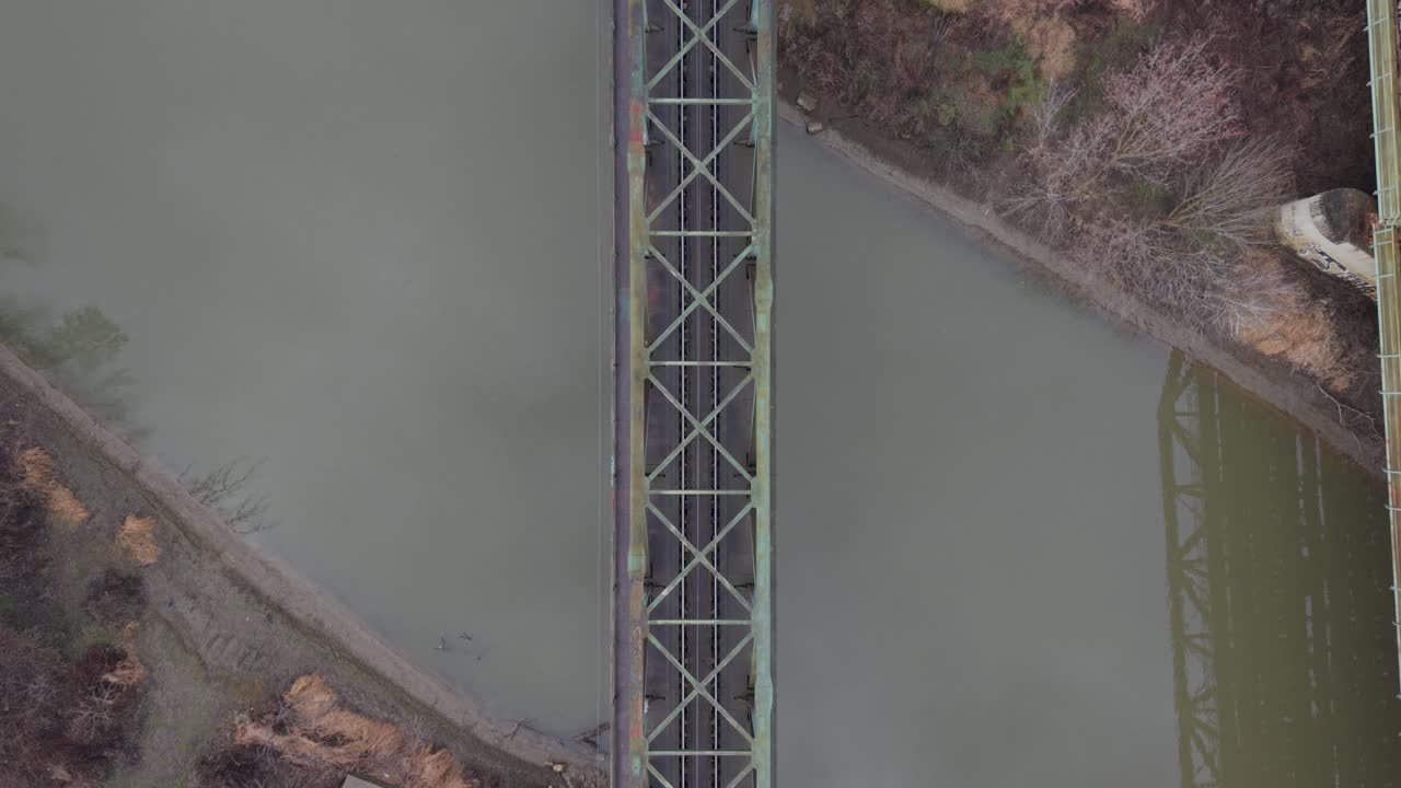 Veduta aerea dall'alto di un ponte ferroviario in acciaio che attraversa un fiume tranquillo. La struttura simmetrica, le tonalità smorzate dell'acqua e la vegetazione invernale enfatizzano l'infrastruttura e l'ingegneria in un paesaggio minimale.