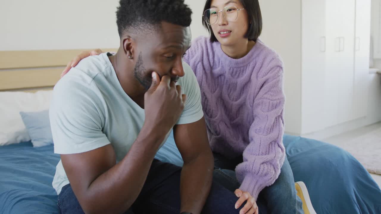 Sad diverse couple sitting on couch and talking in bedroom