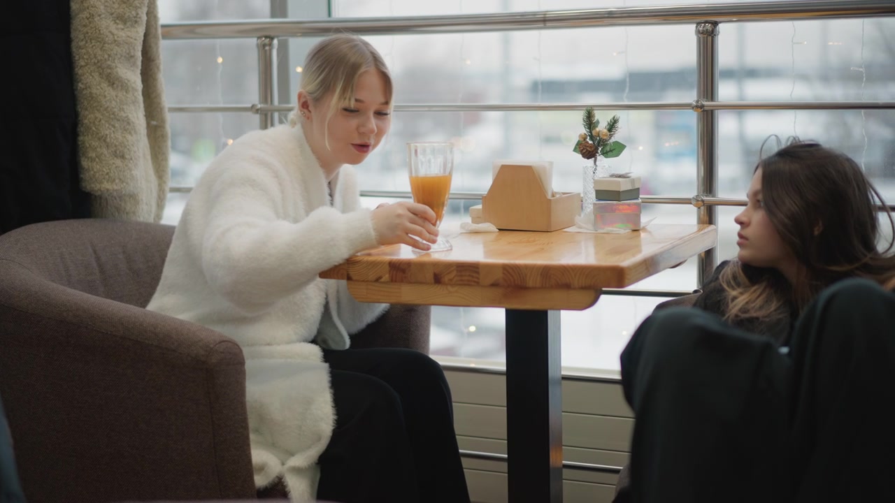Frau in weißem Pullover beugt sich in einer Flughafen-Lounge über einen Holztisch, um eine Essensbox zum Mitnehmen aufzuheben, während sie ein orangefarbenes Getränk hält, was schnelle Bewegung, konzentrierten Ausdruck und eine ungezwungene Reise-Mahlzeit zeigt.