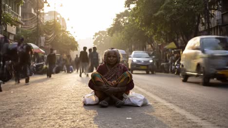 An elderly woman sits on a busy city street with bags by her side as people hurry past her.