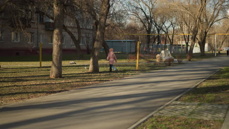 little girl in a pink cap and jacket joyfully chases a pigeon on a sunny park pathway, wearing blue jeans