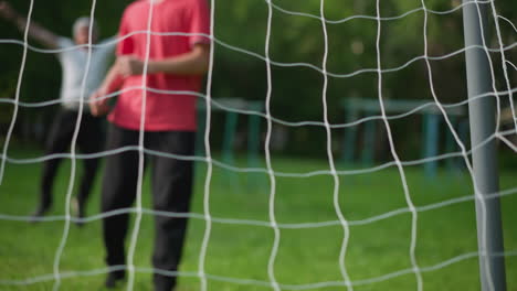 a close-up of a boy in a red top shoots the ball into the net, the goalkeeper misses the save, and a man in the background, blurred, is seen celebrating the goal