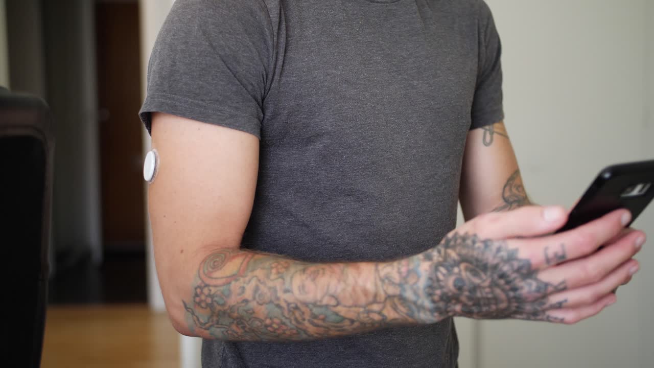 A tattooed young man with diabetes uses his phone to check his blood glucose levels with glucose reading sensor on his arm and then watches the info on the phone
