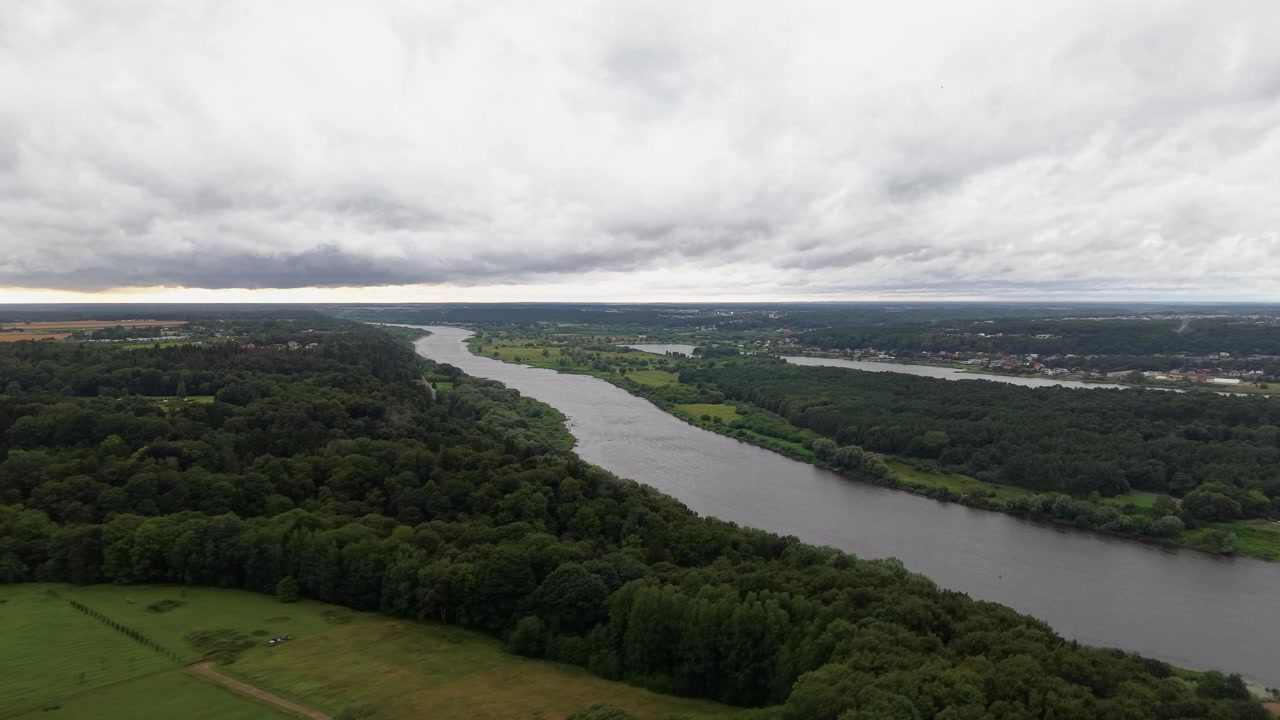 Un video con drone cattura un'ampia veduta aerea di un fiume tranquillo che serpeggia attraverso foreste lussureggianti e campi aperti sotto un cielo cupo e coperto, offrendo uno scenario paesaggistico naturale mozzafiato