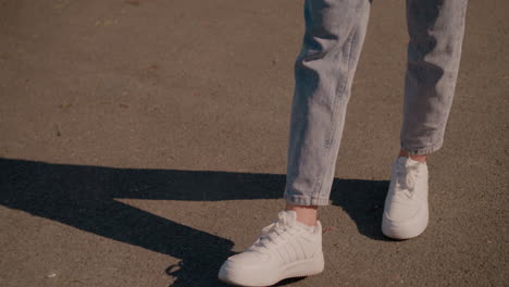 close-up leg view of individual wearing jeans and white sneakers walking on tarred road with cracks around with sunlight casting a long shadow on ground