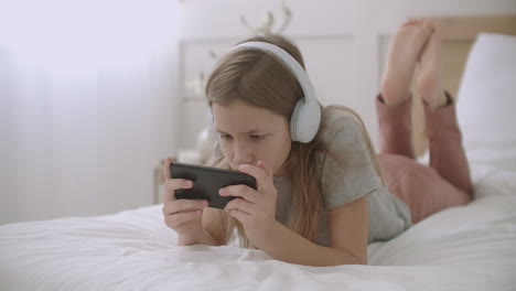 La-Niña-Está-Jugando-En-Un-Teléfono-Inteligente-Con-Internet-Viendo-Imágenes-Y-Fotografías-Deslizando-La-Pantalla-Táctil-Escuchando-Música-Con-Auriculares