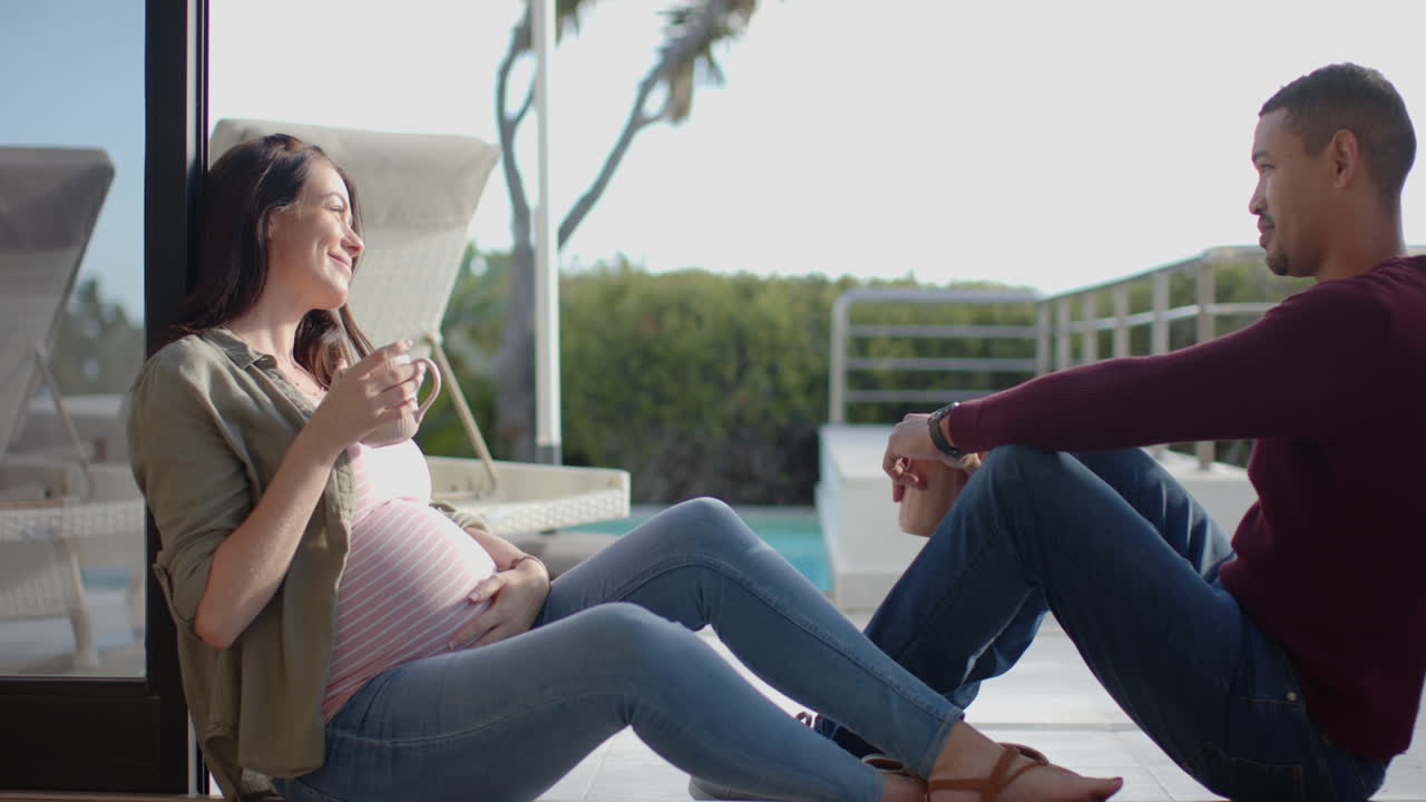 Pregnant woman drinking coffee and sitting with partner on patio, smiling