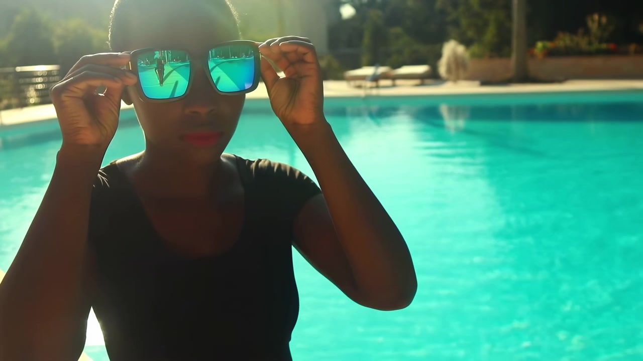 Close-up scene shows person lifting mirrored sunglasses toward face beside pool edge, with turquoise water, sparkling highlights, soft backlight, and relaxed luxury summer mood in bright outdoor