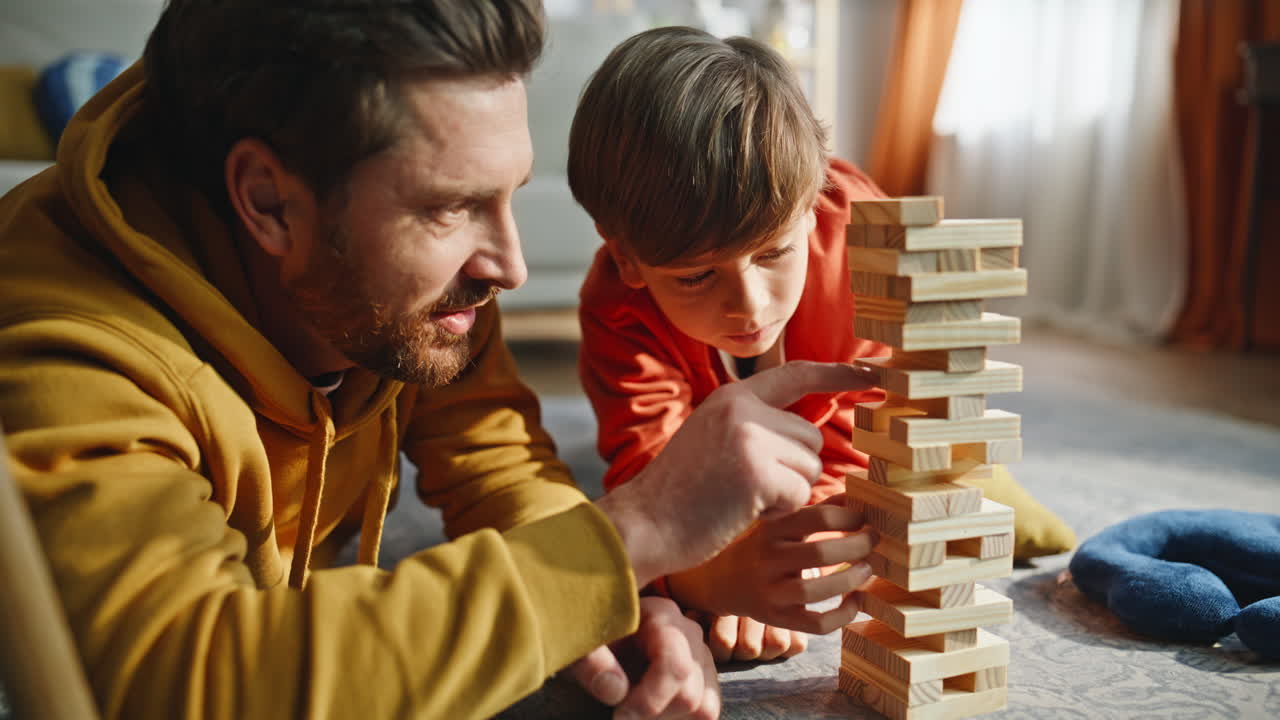 Genitore e figlio si divertono a costruire una torre di legno con Jenga sdraiati sul pavimento del soggiorno, primo piano.