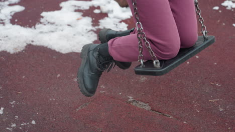 leg view of person kneeling on swing with right knee placed before left, wearing black boots on red textured ground with patches of snow