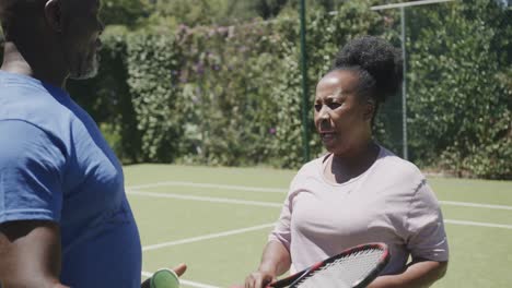 Glückliches-älteres-Afroamerikanisches-Paar-Mit-Tennisschlägern,-Das-Sich-In-Zeitlupe-Auf-Dem-Tennisplatz-Unterhält