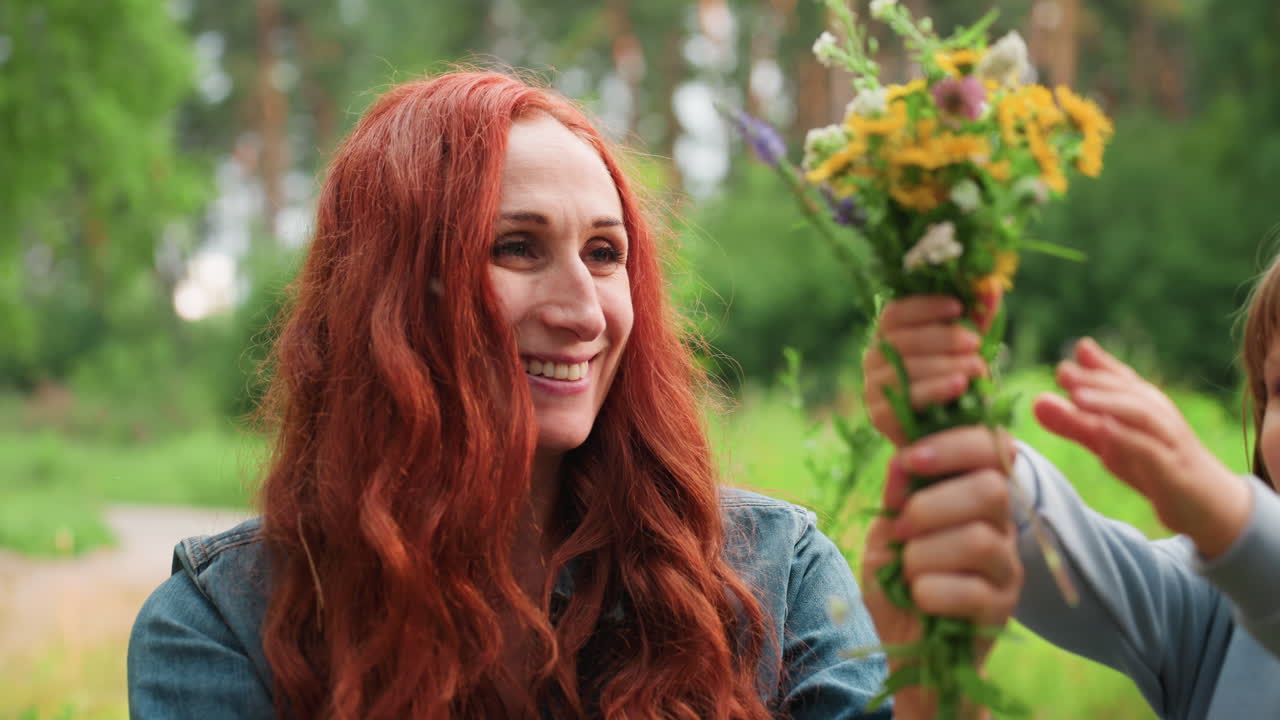 Happy mom joins bouquets picked by family, smiling warmly as she smells fresh wildflowers in sunny forest, showing love, care, and tenderness in peaceful outdoor moment surrounded by green nature