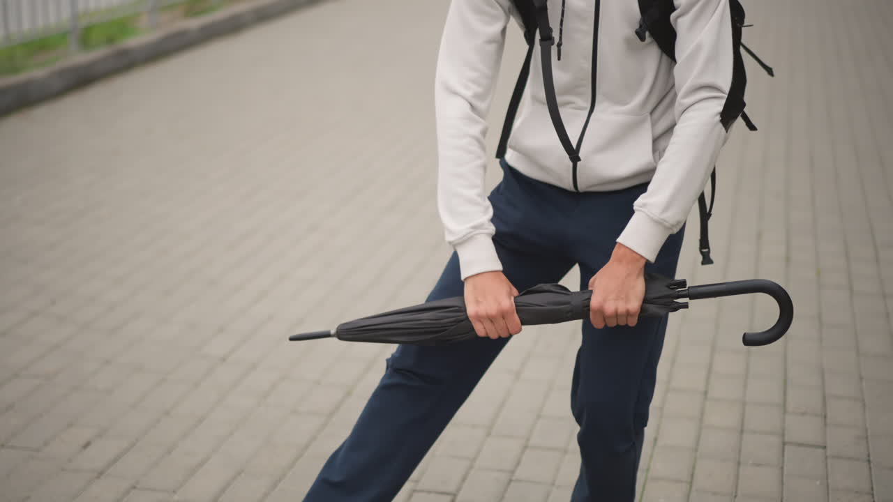 Young man adjusting umbrella on rainy day, Closeup of hands holding umbrella over city sidewalk during rain, Casual young man with backpack adjusting umbrella as it rains on city pavement