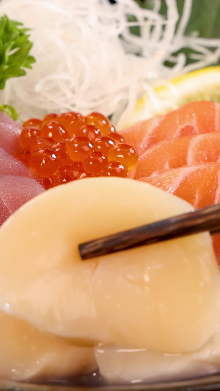 Đũa gắp nhẹ nhàng một miếng sashimi sò điệp từ bộ sushi Nhật Bản đầy màu sắc, cùng với trứng cá hồi, cá ngừ và lát cá hồi dưới ánh sáng đều, rực rỡ.