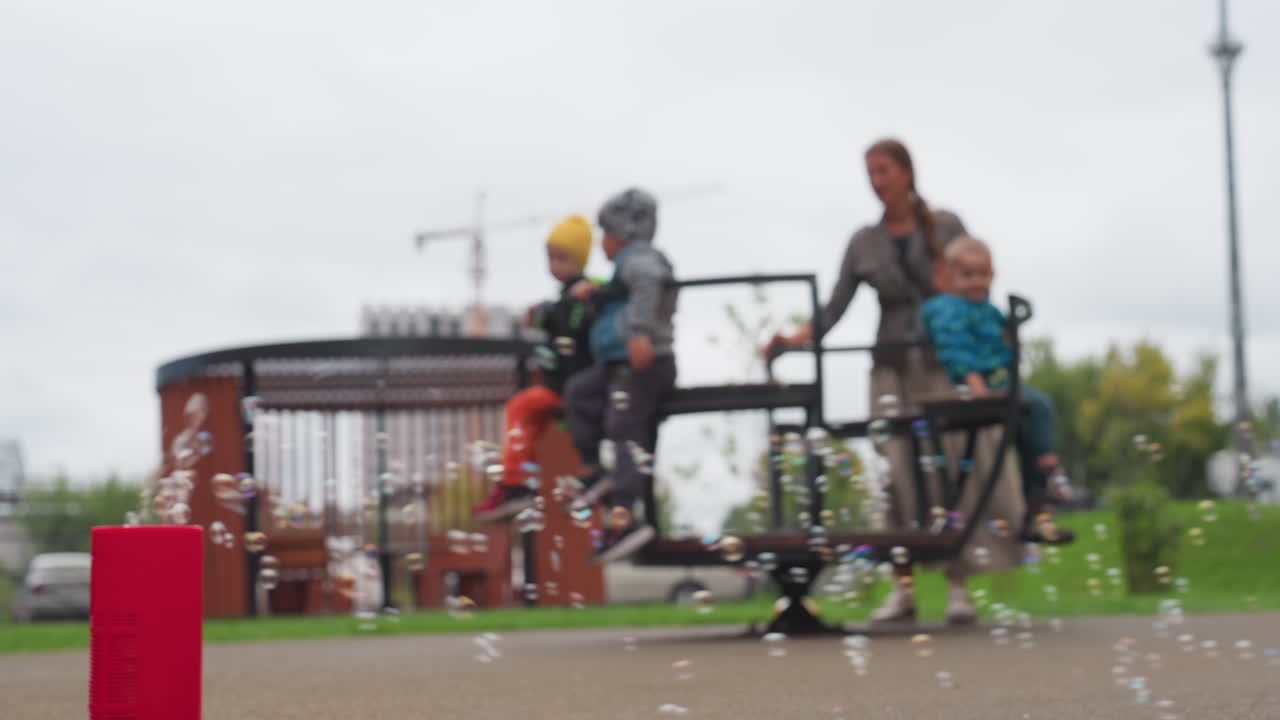 Blur background shows caregiver rotating children on merry go round anticlockwise while red bubble machine blows soap bubbles in foreground, cloudy sky over park, playful spin