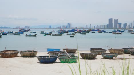 panoramic static view of sandy beach in da nang city vietnam, basket boats rowboat on shore, fishing boats and canoe in sea, cityscape and towers in horizon, traditional local harbor