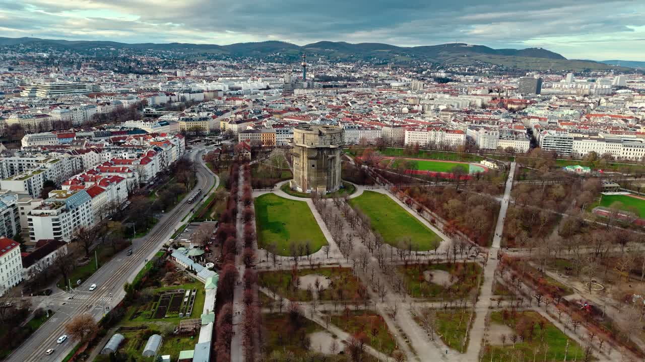 Dronebillede i fugleperspektiv, der panorerer til venstre rundt om Augarten-tårnet i Wien ved solnedgang og viser skyskraberen, den omkringliggende park, de nøgne træer, bygningerne og det varme sollys