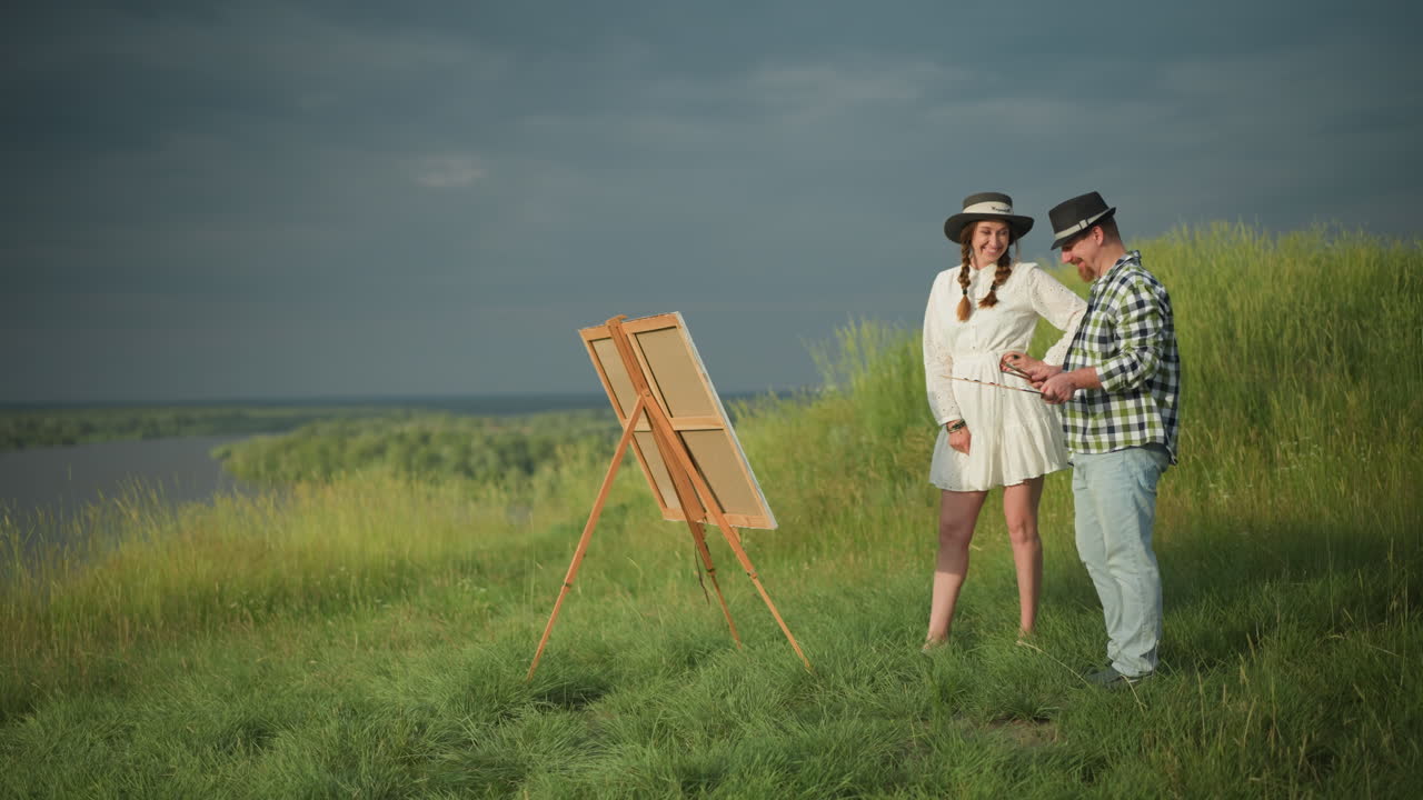 체커드 셔츠와 청바지를 입은 화가 한 명이 팔레트를 들고 하 드레스와 모자를 입은 여자가 그의 에 서서 잔디의 캔버스를 가리키고 있습니다.