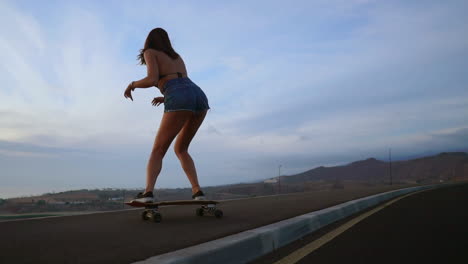 Against-a-backdrop-of-mountains,-a-beautiful-skateboarder-in-shorts-glides-on-her-board-along-a-mountain-road-during-sunset-in-slow-motion