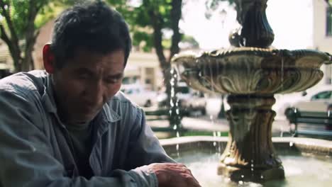 a man sits contemplatively by a fountain in a bustling park, absorbing the vibrant atmosphere and warm sunlight while enjoying the surroundings and people passing by.