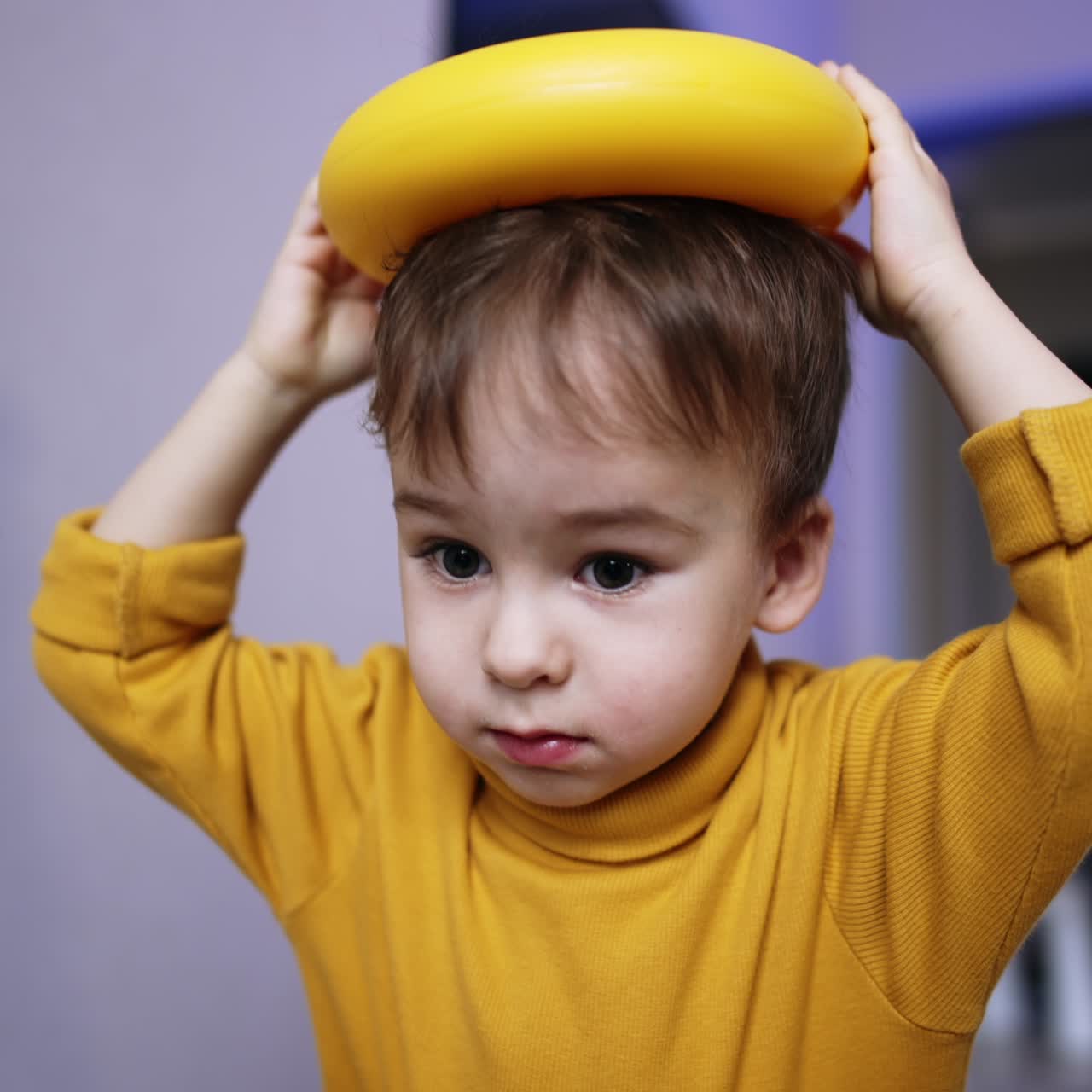 Beautiful brunet male kid in yellow sweater plays with toys. Toddler places ring from toy pyramid on his head and drops it. Close up