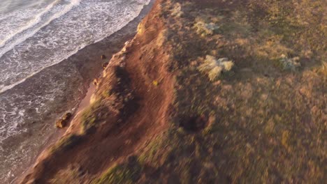 spectacular fpv flight over edge of giant lighting cliffs during sunset - ocean waves reaching sandy beach - mar del plata,argentina