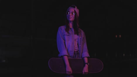 teenager caucasian girl with glasses and cap posing for the camera while holding skateboard outdoors at night