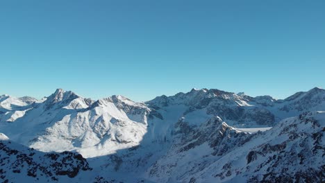 Aerial-view-overlooking-rocky-mountain-ranges,-snowy-winter-landscape---reverse,-drone-shot