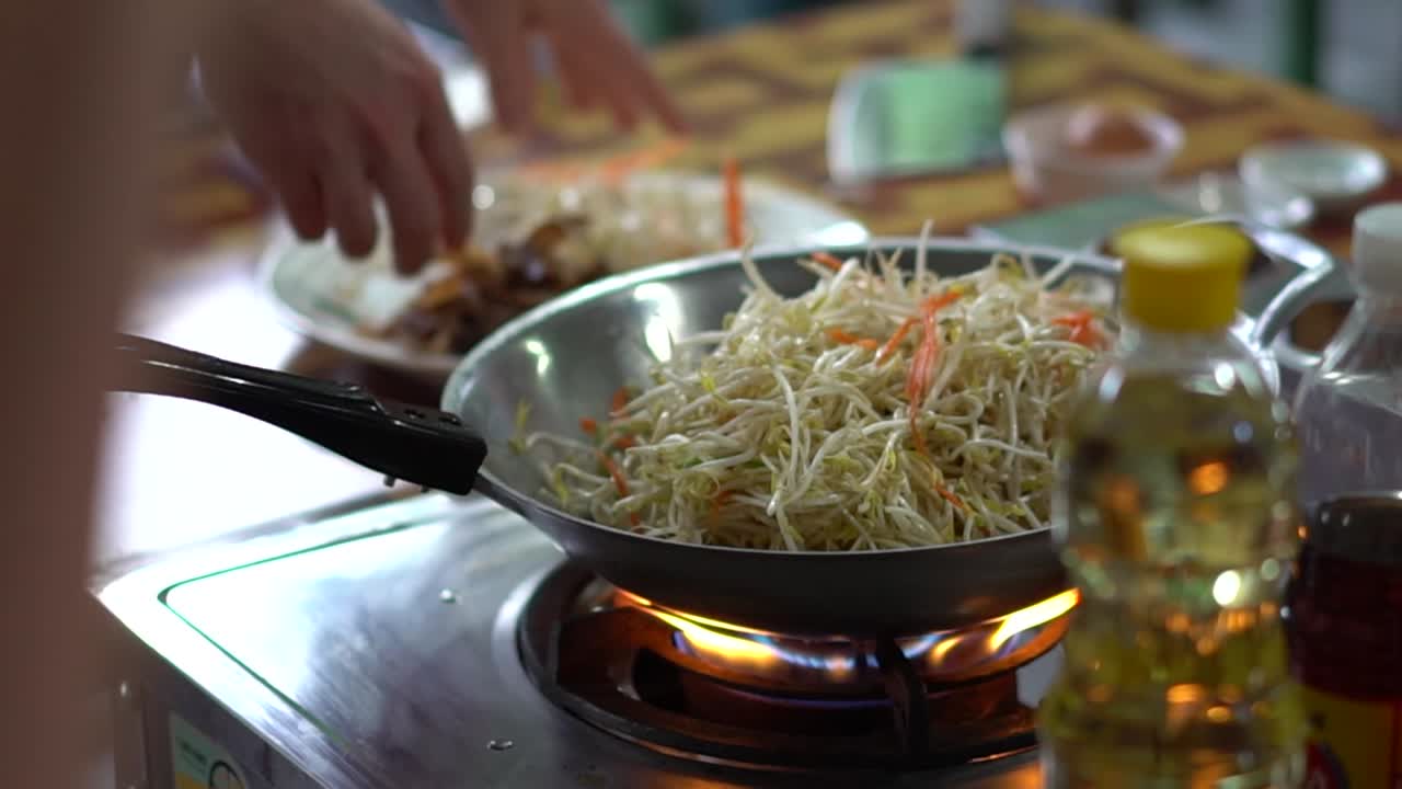 Person lægger håndfulde grøntsager i en wok på en kogeplade ved høj varme for at lave Pad Thai i et udekøkken i Bangkok, Thailand, SLOWMOTION
