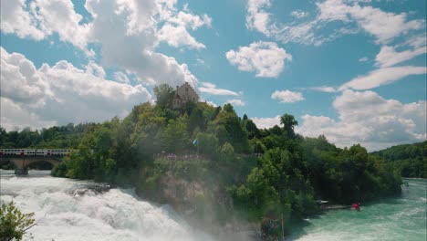 Wide view of the Rhine Falls, with powerful turquoise water rushing over the rocks. Historic building stands on the cliff above, while tourists gather on viewing platforms for a closer experience