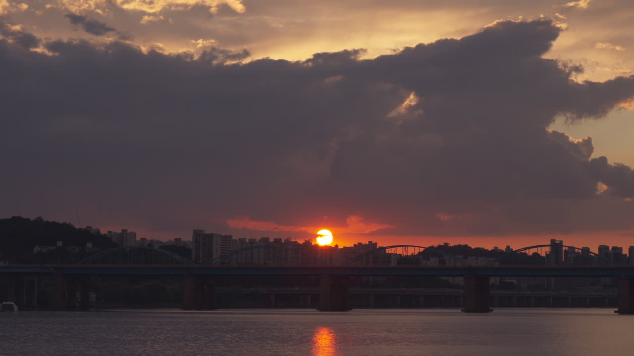 Sunset over Seoul’s Dongjak Bridge with the Han River glowing below. High-rise apartments silhouette against the fiery sky as the sun dips, An airplane can be seen passing by the glowing sun